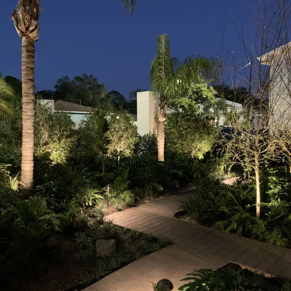 Uplit palms and low-voltage path lights along a boardwalk through tropical beds at dusk