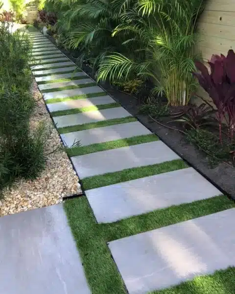 Large square pavers set in a strip of green turf along a side yard, with palms and red ti plants beside them