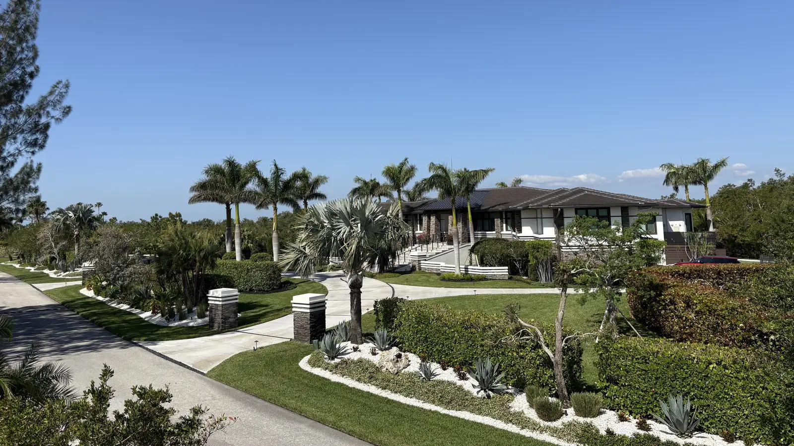 Bands of green lawn curving between white river rock beds, clipped hedges and tall palms in front of a large home